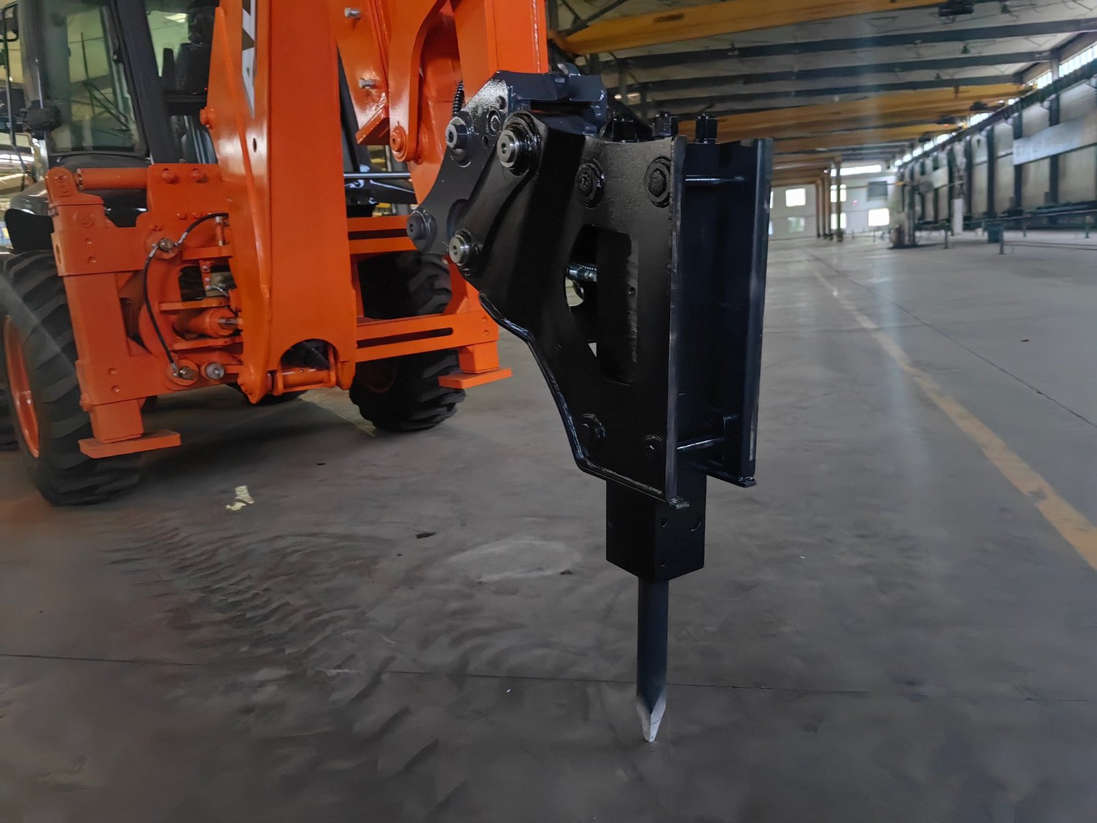 A hydraulic breaker mounted on the backhoe arm of an orange backhoe loader, standing on the workshop floor