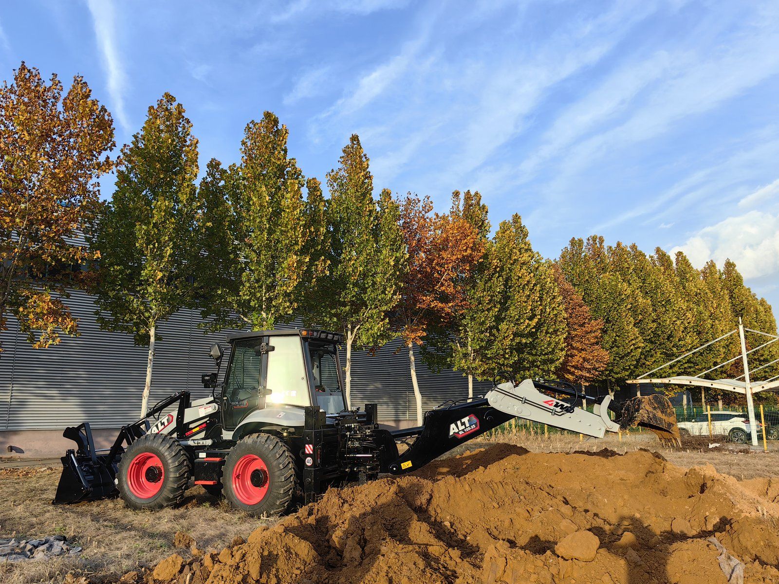 A backhoe loader working a mound of soil on a customer site, its backhoe arm extended over the spoil heap