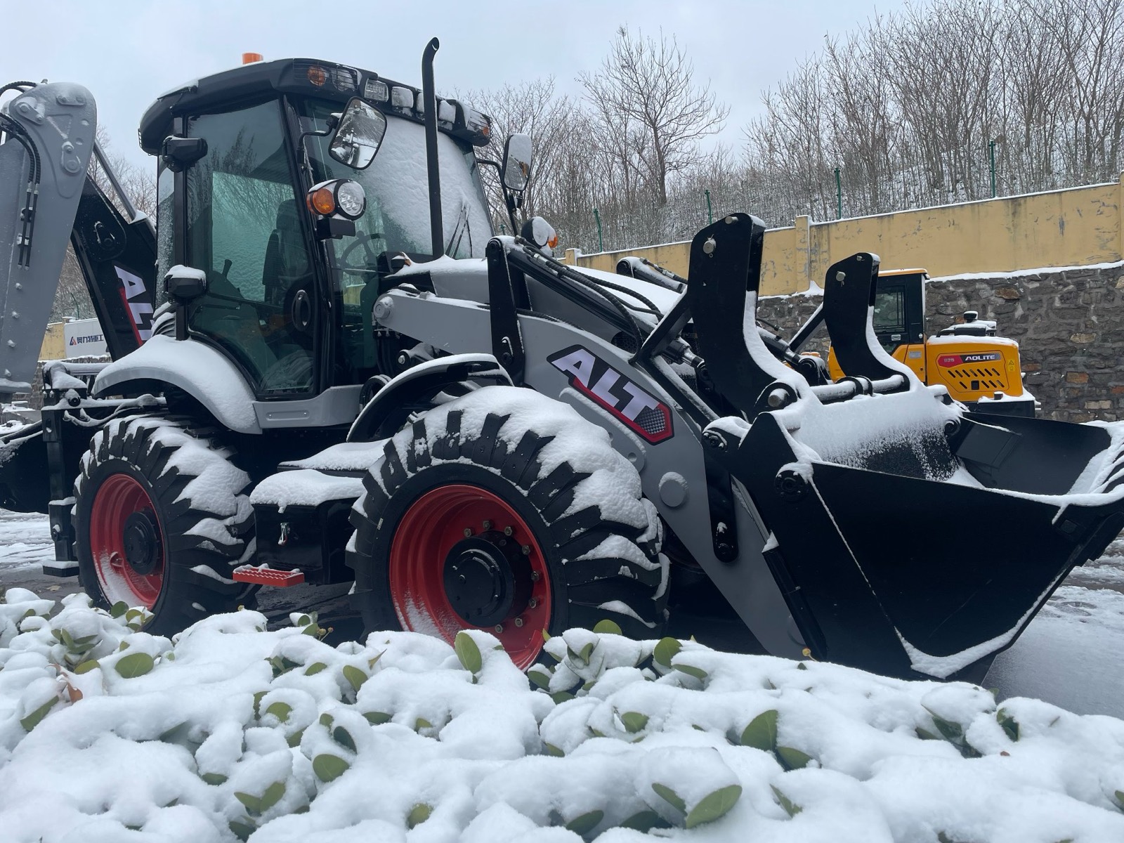 An ALT backhoe loader standing in a snow-covered yard with its front bucket full of snow, a second AOLITE machine parked behind it