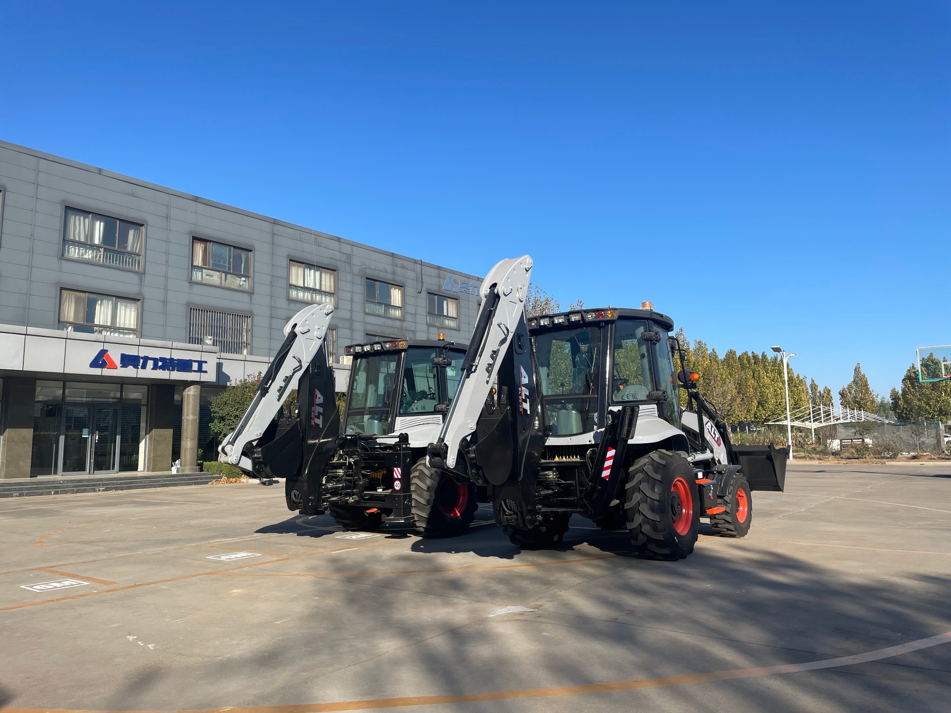 A row of AOLITE backhoe loaders parked in the plant yard in front of the factory building on a clear day