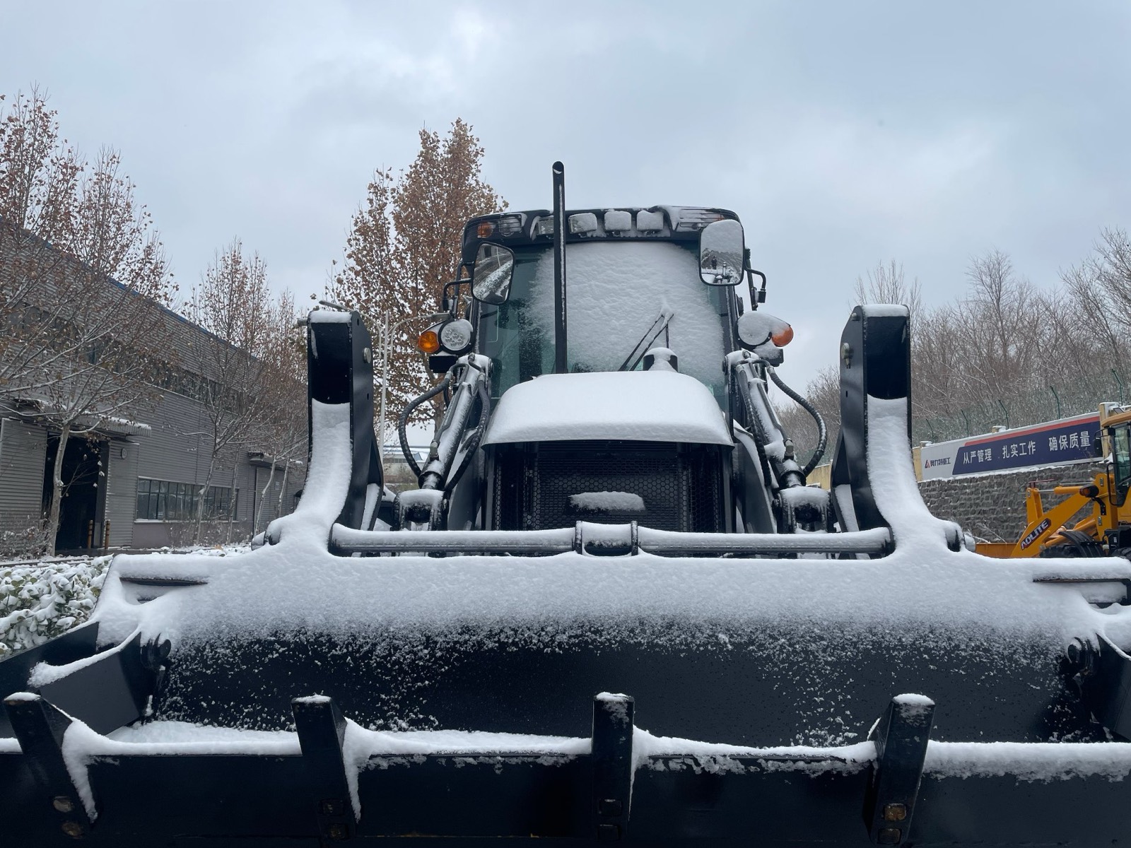 Front view of a backhoe loader covered in snow, with snow sitting on the loader arms, bucket, mirrors and cab roof
