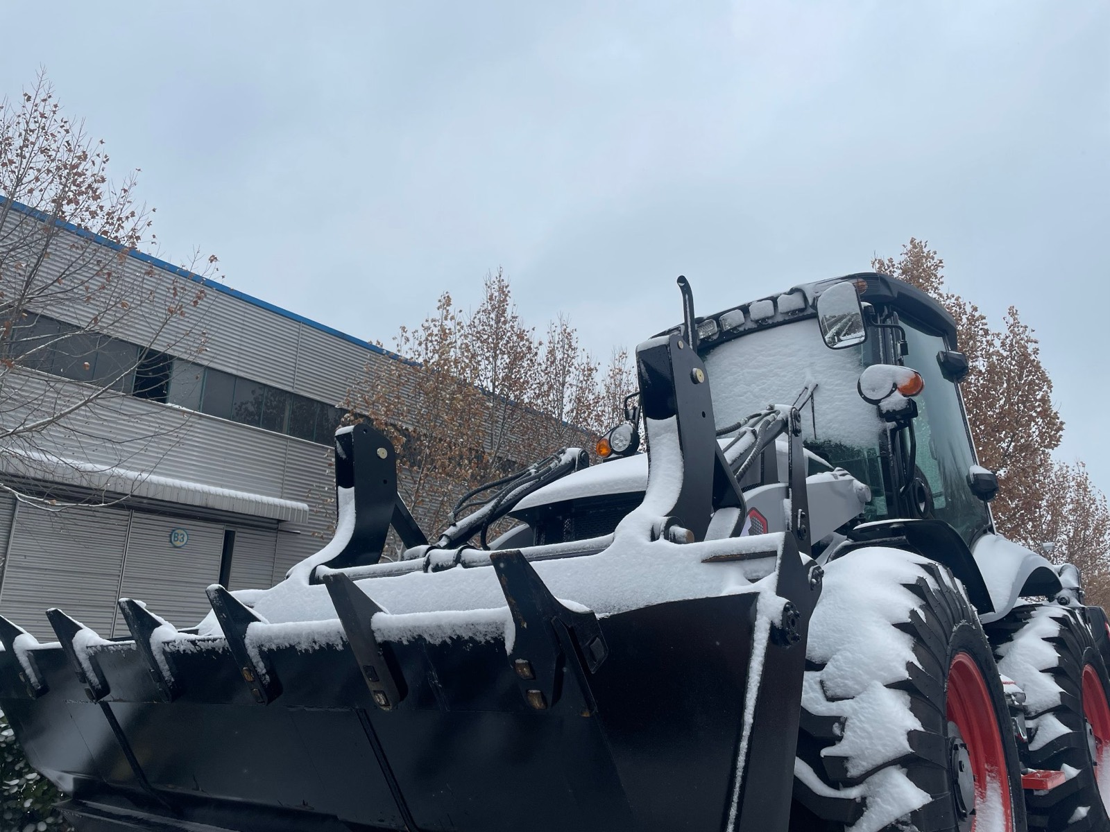 A backhoe loader standing in snow in the factory yard, fresh snow covering the loader bucket, arms and cab roof