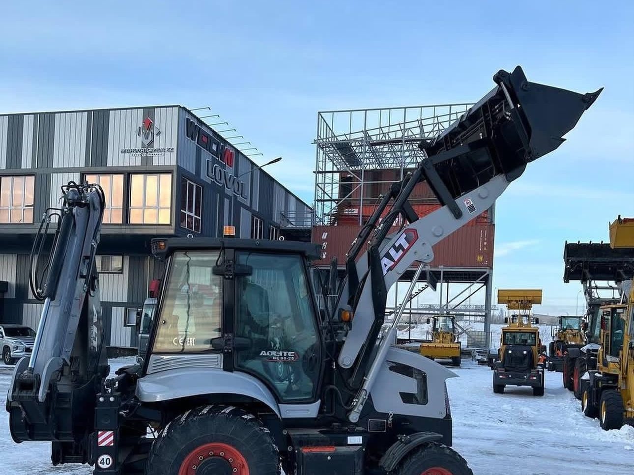 Backhoe loader parked in a snow-covered yard with a row of machines waiting behind it