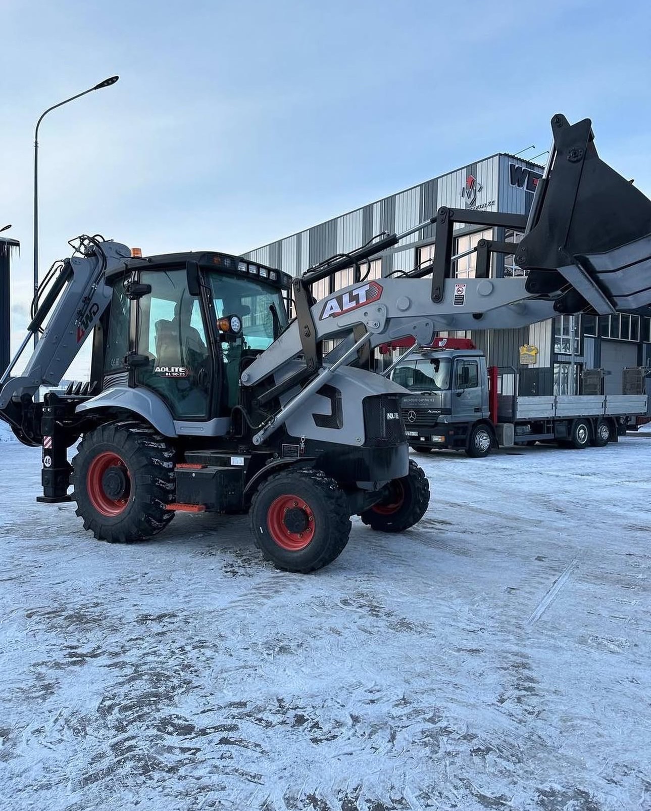 An AOLITE backhoe loader with the loader bucket raised, parked on frozen snow-covered ground with a flatbed truck in the background