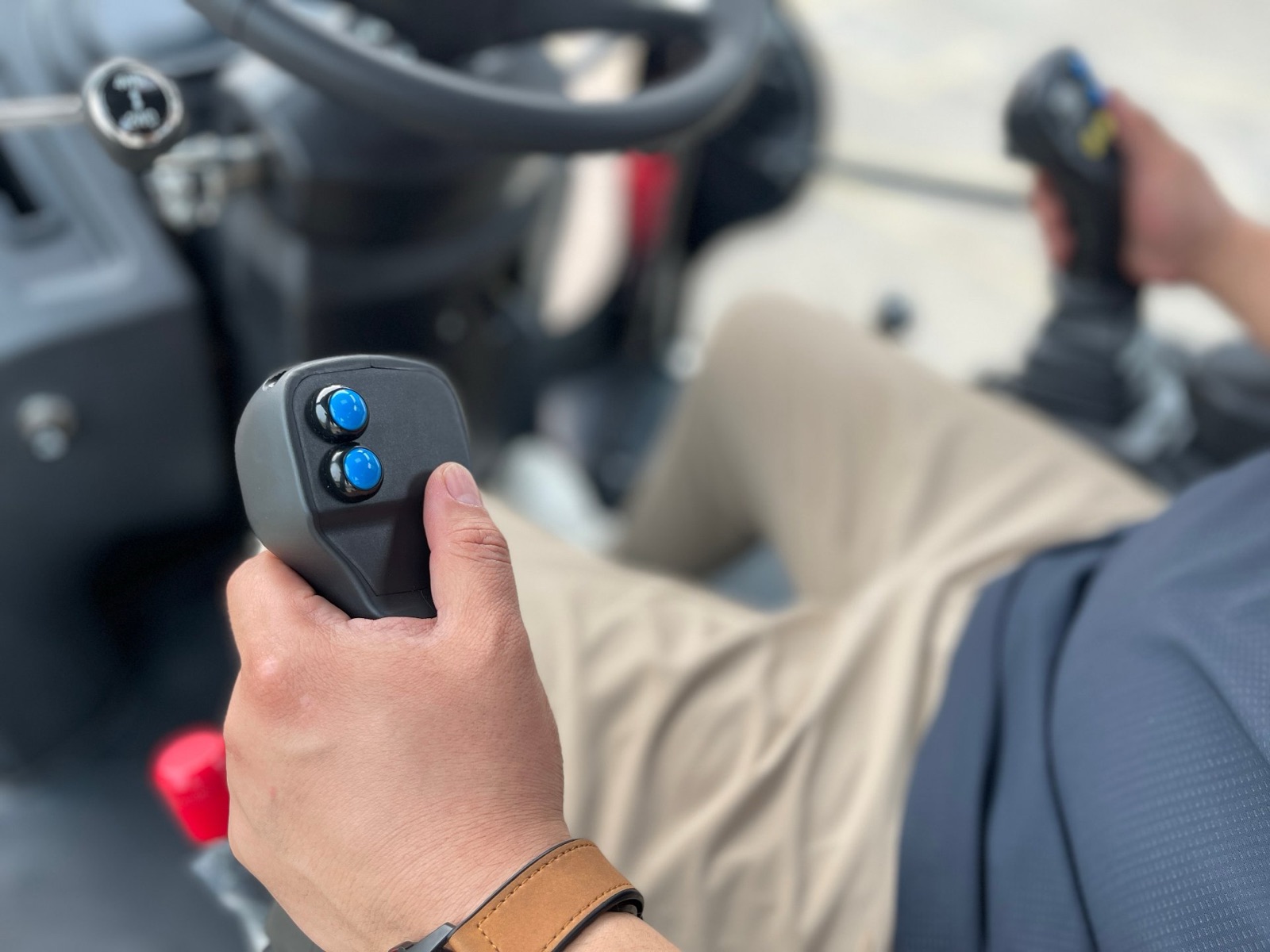 A hand gripping the pilot control joystick with two blue buttons inside the cab of a backhoe loader, a second joystick in the operator's other hand
