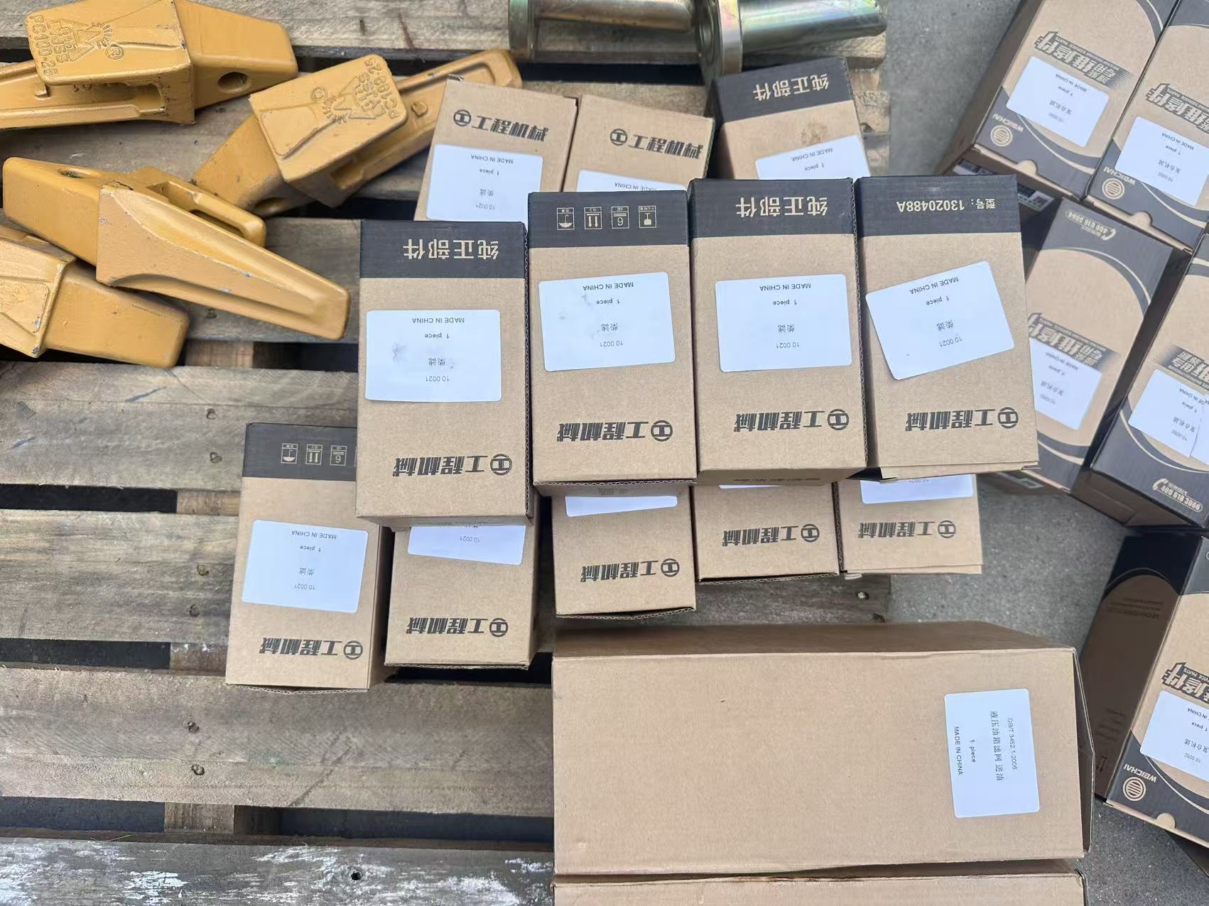 Backhoe loader bucket teeth and labelled genuine-part boxes with part numbers on a wooden pallet
